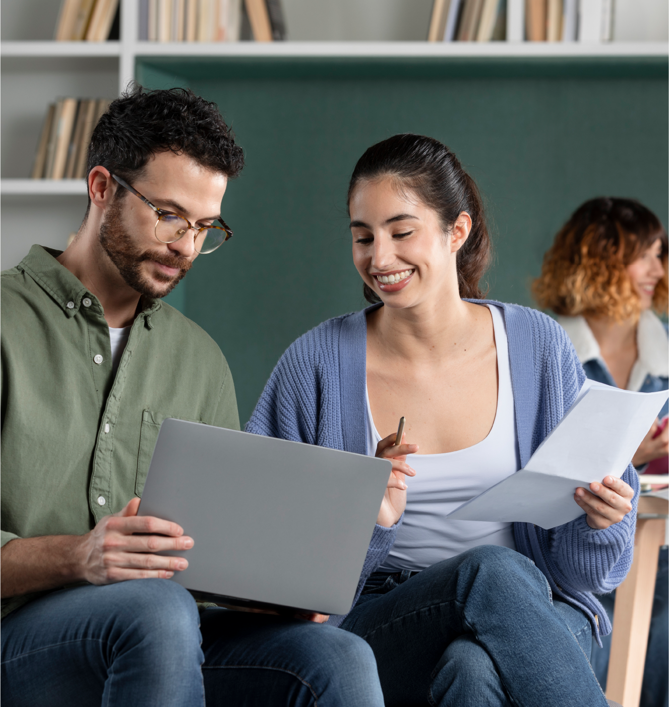Um homem com um laptop no colo e uma mulher com um papel e lápis na mão sentados lado a lado, felizes, discutindo sobre algo