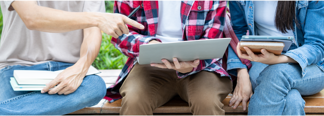 close-up nas mãos de três pessoas sentadas lado a lado com materiais de pesquisas, laptop, livros e relatórios, mostrando uns aos outros